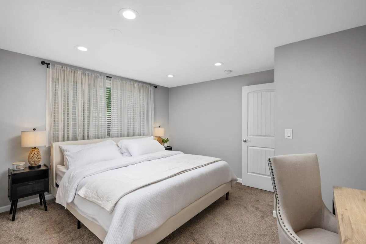 A serene bedroom featuring a comfortable bed adorned with crisp linens, a nightstand with a lamp, a desk and chair in the corner, and soft curtains framing the window. The space feels uncluttered and restful, perfect for both sleeping and working.