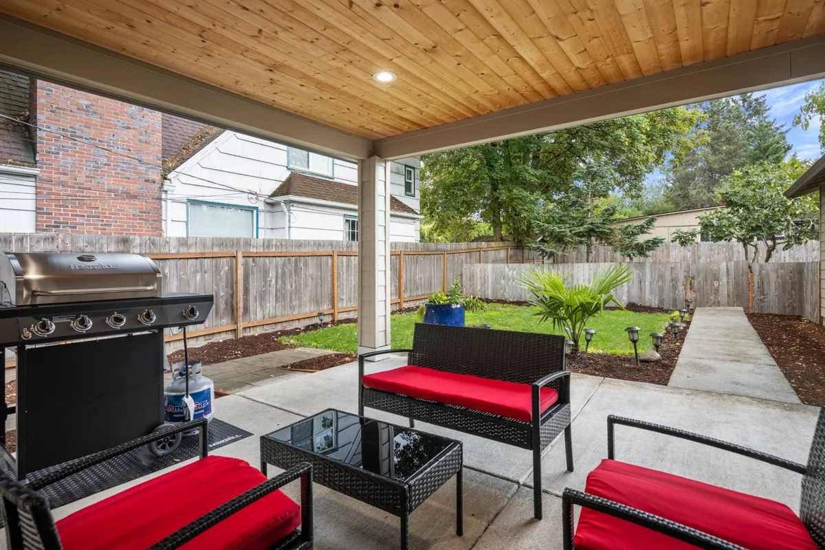 This image showcases an inviting backyard scene with an outdoor grill, comfortable seating, and a lush garden area. The setting clearly depicts a fenced space ideal for relaxing, entertaining guests, or sharing meals with family and friends. The visible perimeter gives a sense of privacy and security, while the mix of greenery and furnishings invites both leisure and play.