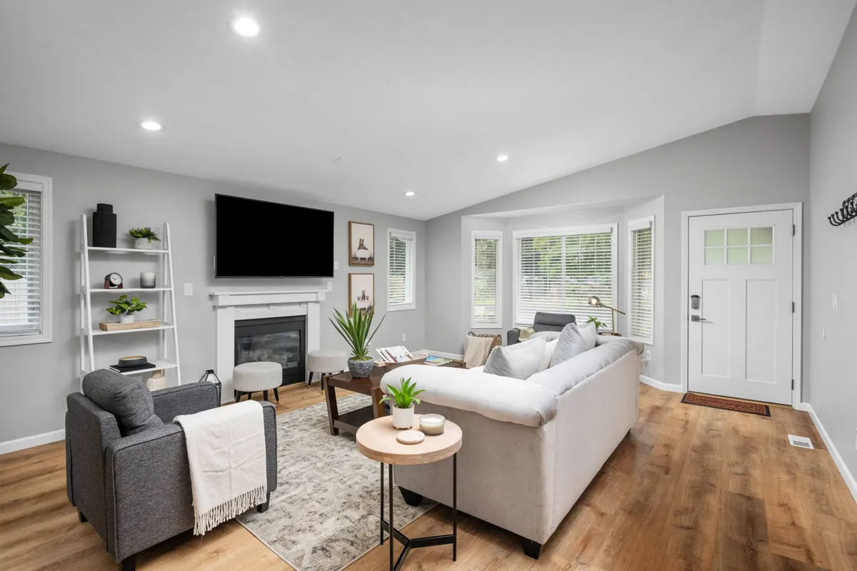 A cozy living room with a fireplace, comfortable sofa, coffee table, window seating, and a bookshelf. Large windows let in streams of natural light, enhancing the inviting and intimate ambiance of the space.