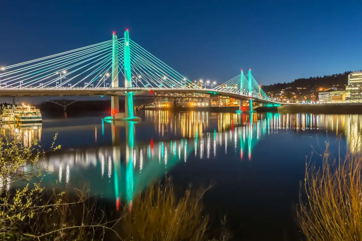 An illuminated city bridge stretches over a calm river, with reflections shimmering on the water and the city skyline glowing in the background. The scene is tranquil yet lively, capturing the spirit of evening explorations.