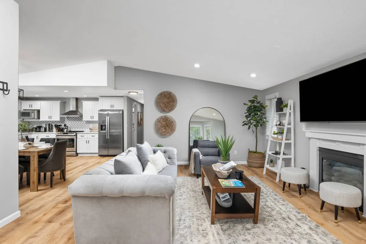 A spacious, sunlit open-plan interior featuring a large sectional sofa, dining area with a wooden table, modern kitchen appliances, and a fireplace with a TV above. Decorative wall mirrors enhance the brightness and sense of space. The room is filled with natural light, creating an inviting and comfortable setting.